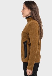 Leichte Fleecejacke in dunklem Amber mit hohem Kragen, seitlichem Reißverschluss und schwarzen Akzenten; strukturierter Stoff mit figurnahem Design.