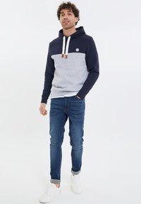 Kapuzensweatshirt in Marineblau und Grau, mit einem Farbblockdesign, weißen Kordeln und braunen Lederelementen. Getragen mit engen Jeans und Turnschuhen.