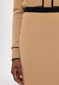 Beige knit dress featuring a black ribbed waistband and cuff details; soft texture; close-up of sleeve and hand on fabric surface.