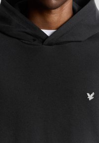 Sort hættetrøje lavet af tekstureret stof. Har en kengurulomme, snoretræk til hætten og et lille sølvlogo på brystet. Simple design.