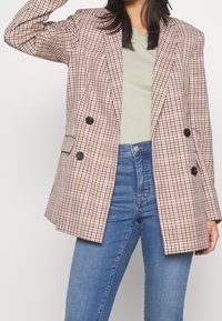 Dubbelknäppt blazer i en beige och mörkbrun rutad mönstring. Har svarta knappar, strukturerade axlar och en avslappnad passform. Bärs över en ljusgrön topp.