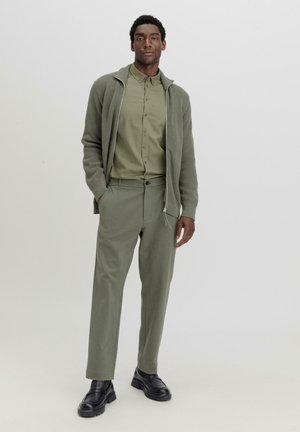 Olijfgroene ritssluiting trui met ribbeltextuur over een bijpassende button-up shirt, gecombineerd met bijpassende broeken en zwarte instappers.