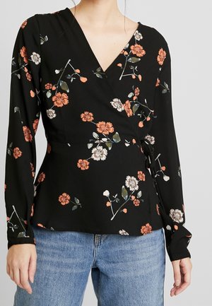 Sort langærmet slå-om-bluse med orange og hvidt blomsterprint, bundet i taljen, båret med lyseblå denimjeans.