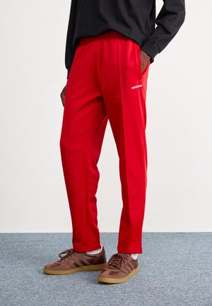Persona con pantalones deportivos Adidas de color rojo brillante y zapatillas marrones, de pie sobre una alfombra gris contra un fondo blanco.