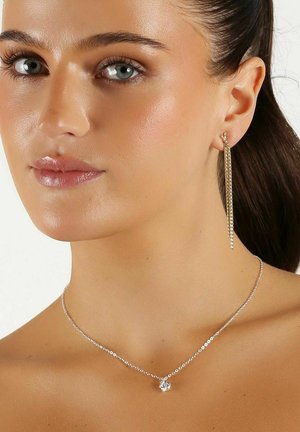 Silver chainhalsband med en central berlock, tillsammans med en guldig hängörhänge med lång kedjedetalj. Hudtonen är naturlig.