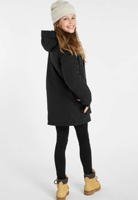 Zwart gewatteerd jack met een capuchon, gedragen over zwarte leggings. Afgewerkt met een beige beanie en gele laarzen. Eenvoudig en functioneel ontwerp.