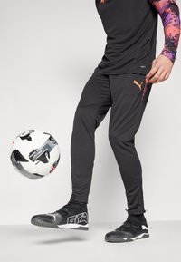 Schwarze Sporthosen mit einem orangefarbenen Logo und einem passenden schwarz-weißen Fußball, kombiniert mit schwarzen Sportschuhen mit einem weißen Akzent.