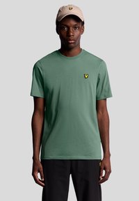T-shirt verde in cotone a maniche corte, con un piccolo logo giallo ricamato sul lato sinistro del petto. Abbinato a un cappellino beige.