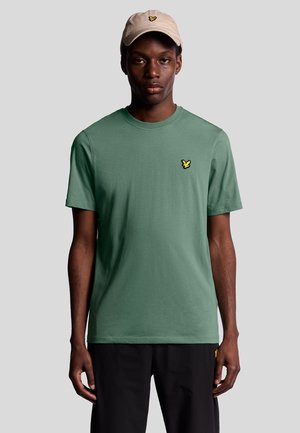 Green cotton t-shirt with short sleeves, featuring a small yellow embroidered logo on the left chest. Paired with a beige cap.