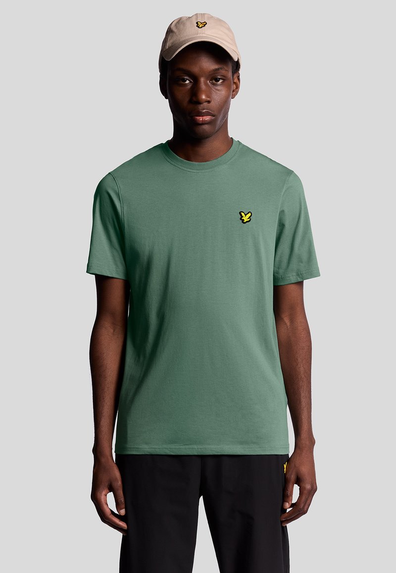 T-shirt verde in cotone a maniche corte, con un piccolo logo giallo ricamato sul lato sinistro del petto. Abbinato a un cappellino beige.