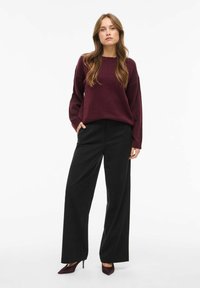 Frau mit langen Haaren, die einen lockeren, dunkelroten Pullover, schwarze weit geschnittene Hosen und dunkelrote High Heels trägt, steht mit den Händen in den Taschen.