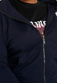 Námořnický zipový svetr s dlouhými rukávy, vyrobený z měkkého bavlněného materiálu, s předními kapsami a hladkým zipem. Nositý přes černé tričko s potiskem.