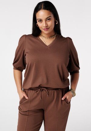 Femme aux cheveux lisses et foncés portant un haut marron à encolure en V avec des manches bouffantes et un pantalon assorti, accessoirisée de bijoux en or.