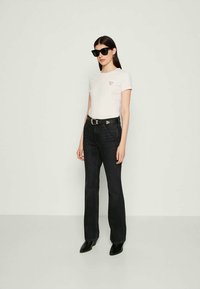 Woman wearing black sunglasses, white fitted short-sleeve t-shirt with logo, black high-waisted jeans, black belt, and black pointed shoes standing.