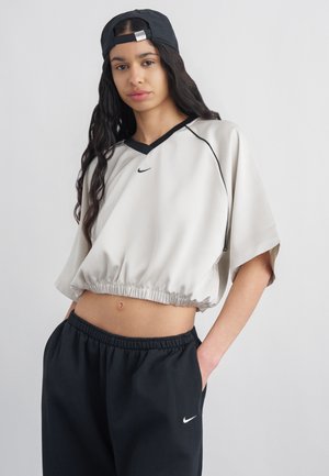 Νέος/νέα φοράει μαύρο καπέλο φορεμένο ανάποδα, ανοιχτό cropped πουκάμισο και μαύρο φόρμα με τα χέρια στις τσέπες, μπροστά σε απλό φόντο.