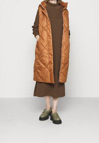 Tikkivuorattu kuparinvärinen toppaliivi hupulla, jossa kaksi sivutaskua. Päällä ruskean ribbineulosmekon päällä ja yhdistetty vihreisiin puukenkäihin vaalealla taustalla.