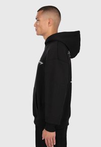 Męski model w czarnej bluzie z kapturem i kieszenią z przodu, stojący w profilu na jednolitym jasnoszarym tle.