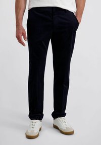 Man in een donkerblauwe corduroy broek en witte sneakers met gomzolen, staand met één hand in zijn zak tegen een effen achtergrond.