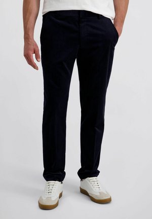Man in een donkerblauwe corduroy broek en witte sneakers met gomzolen, staand met één hand in zijn zak tegen een effen achtergrond.