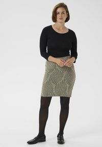 Schwarzes Langarmtop, kombiniert mit einem taillierten knielangen Rock mit beigem Grundton und schwarzen wellenförmigen Mustern, ergänzt durch schwarze Strumpfhosen und Loafers.