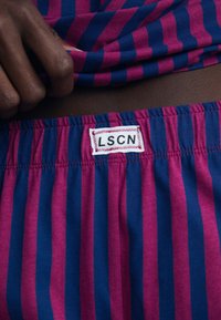Randiga tygshorts med växlande rosa och blå ränder, med en midjeresår som har en etikett som säger "LSCN." Mjuk textur och elastisk passform.