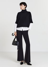Schwarzer, gestrickter Rollkragenpullover mit kurzen Ärmeln, über einem weißen Hemd getragen. Passende schwarze Schlaghosen und eine kleine schwarze Handtasche runden den Look ab.