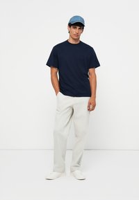 Marineblauw katoenen T-shirt met ronde hals, gecombineerd met lichtgrijze broeken en witte sneakers; draagt een lichtblauwe pet.