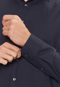 Camicia texturizzata blu scuro con maniche lunghe, polsini con bottoni e un logo discreto. Presenta un colletto e bottoni abbinati.