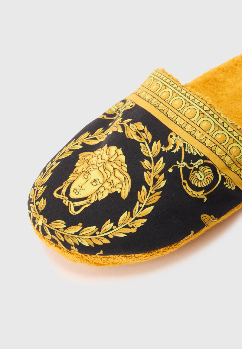 Black and gold ornate slipper with Medusa head emblem, laurel wreath design, and mustard-yellow plush lining on a white background.