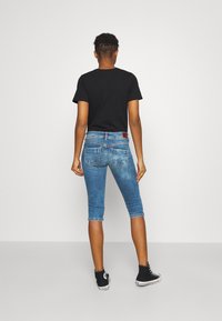 Niebieskie dżinsowe jeansy capri o jasnym kolorze, pięć kieszeni i metka z logo z tyłu. Na sobie czarny t-shirt z krótkim rękawem i sneakersy.