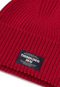 Berretto a coste rosso con un'etichetta nera che recita "Thinking Mu" e ulteriore testo in piccolo riguardante il design a minimo impatto.