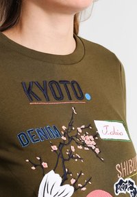 Primer plano de una persona con una camisa verde oliva que tiene bordadas las palabras "KYOTO", "DENIM", "Tokio" y diseños de ramas florales.