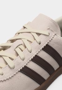 Close-up van een beige suède sneaker met crèmewitte veters en drie donkerbruine strepen met zigzagrand langs de zijkant.