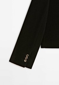 Manche de blazer noir avec trois boutons ronds métalliques disposés verticalement près du poignet sur fond blanc.