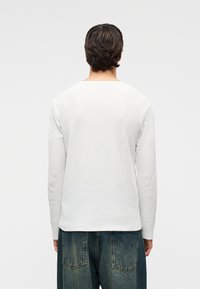 Λευκό πουκάμισο με μακριά μανίκια από υφή υφάσματος, με φαρδύ ντεκολτέ και ελαφρώς κυρτό στρίφωμα, σε συνδυασμό με σκουρόχρωμο τζιν.