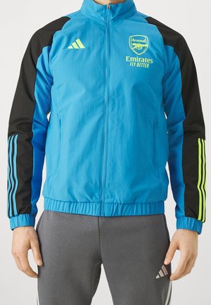 Blau-schwarze Sportjacke mit Reißverschluss, Arsenal-Logo und dem Schriftzug "Emirates Fly Better", getragen mit grauer Adidas-Hose.