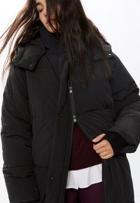 Crna puffer jakna s kapuljačom, sa zip zatvaranjem i dugmadima na preklapanje, kviltanim uzorkom i rebrastim manžetama. Nosi se preko bordo majice.