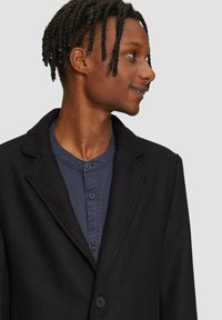 Schwarzer Blazer aus strukturiertem Stoff, mit Reverskragen und Knopfverschluss, kombiniert mit einem marineblauen Hemd mit Rundkragen und Knöpfen.