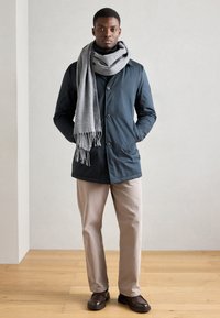 Parka azul-marinho com botões, bolsos laterais e um cachecol cinza com franjas, combinado com calças beje claro e sapatos castanhos escuros, sobre um chão de madeira.