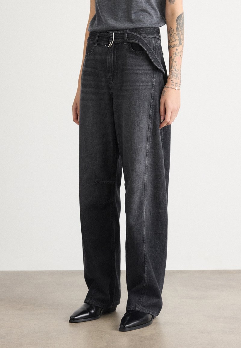 Personne portant un jean noir ample avec un détail de fermeture éclair déboutonné à la taille, assorti à des chaussures noires pointues et une chemise grise.