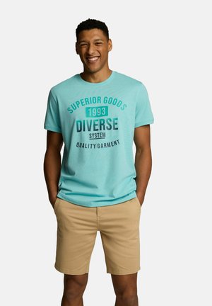 Jasnoniebieska koszulka z krótkim rękawem z ciemnym nadrukiem na przodzie. W połączeniu z beżowymi szortami, model stoi z rękami w kieszeniach, uśmiechając się.