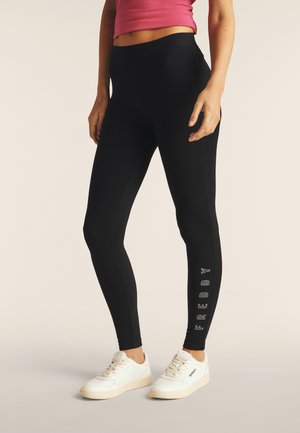 Leggings neri realizzati in tessuto elasticizzato, con vita alta e un logo verticale "FREDDY" in bianco sulla parte inferiore della gamba. Indossati con sneakers bianche.