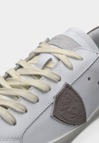 Baskets blanches avec lacets crème, patch logo en daim gris sur le côté et coutures visibles sur la tige en cuir.