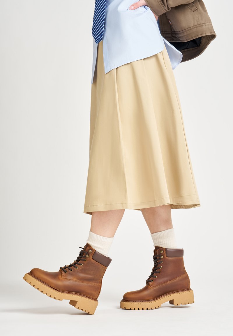 Stivaletti in pelle marrone con suola robusta, caratterizzati da un design con lacci e accenti marroni a contrasto, abbinati a una gonna beige e calzini bianchi.