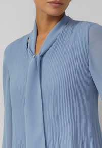 Hellblaue Bluse aus glattem Stoff mit vertikalen Nadelstreifen und einer gebundenen Halsdetail. Lange Ärmel und lockere Passform.