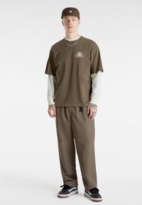 Bruine T-shirt met bergafbeelding, witte lange mouw onderlaag, bruine relaxte broek en zwarte sneakers met witte accenten.
