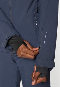 Giacca blu navy con una texture liscia, presenta una tasca con zip sul braccio e polsini elasticizzati. Il logo e i dettagli del materiale sono visibili.