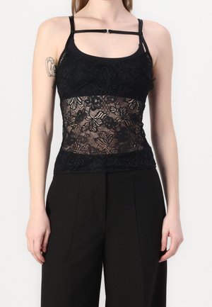 Femme portant un haut sans manches en dentelle noire avec un motif floral et une fine bretelle sur la partie supérieure de la poitrine, associé à un pantalon noir.