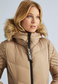 Blond naine, kes kannab beeži puffer-jakki, mille kapuuts on kaunistatud karvaga, kinnitatud musta tõmblukuga, millel on kiri "JACKIT", seisab paremale poole.