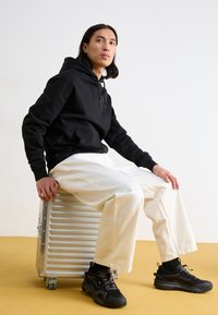 Felpa nera con cappuccio e tasca frontale, abbinata a pantaloni bianchi larghi e sneakers nere spesse, seduto su una custodia metallica argentea.